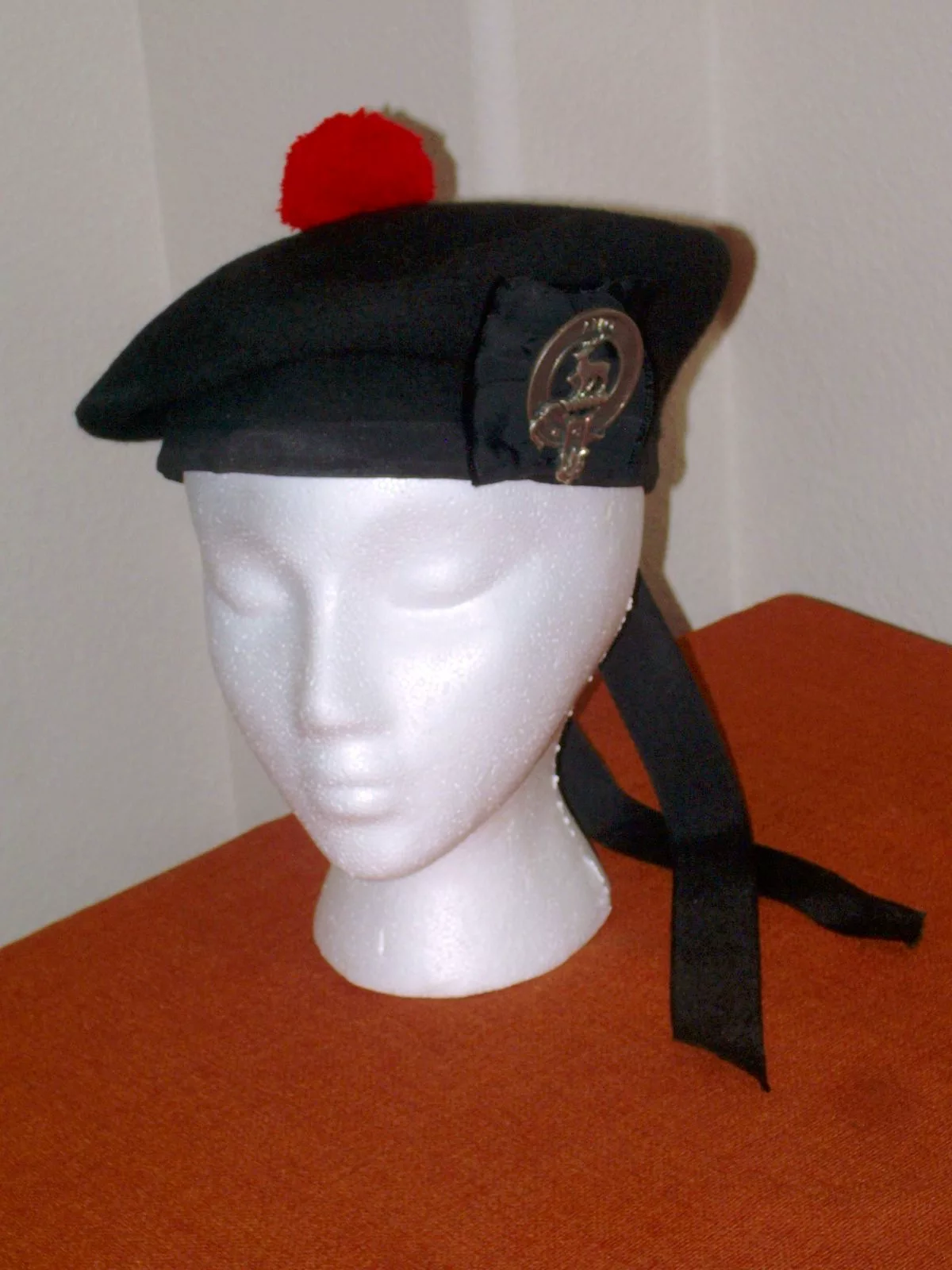 A black wool Balmoral bonnet with a red pom-pom, ribbon tails and a metal clan crest badge, shown on a white foam head