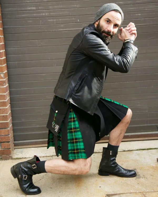 A bearded man kneeling in a black kilt with a green and yellow tartan inner pleat and a large side pocket, worn with a leather jacket and boots