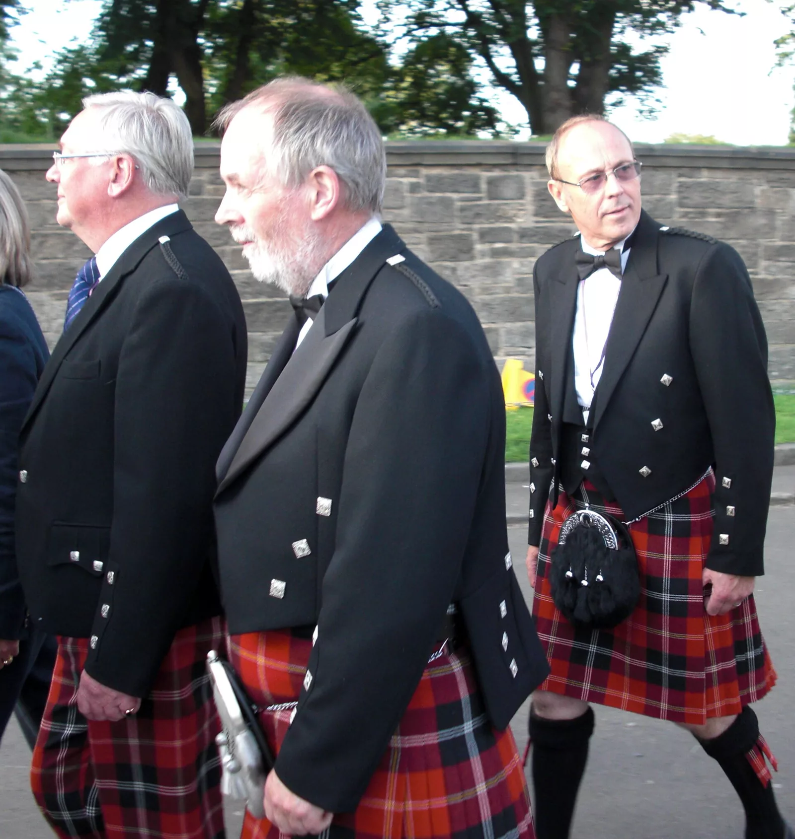 Three men in black jackets and kilts of the same red, black and white tartan, walking along a street