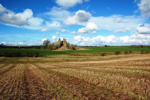 A small ruined stone tower on a low mound beside a farm, seen across a ploughed field under a blue sky.