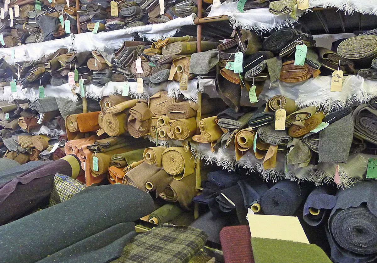 Shelves of rolled and folded woollen cloth in many colours, each bolt with a paper tag, in a cloth warehouse.