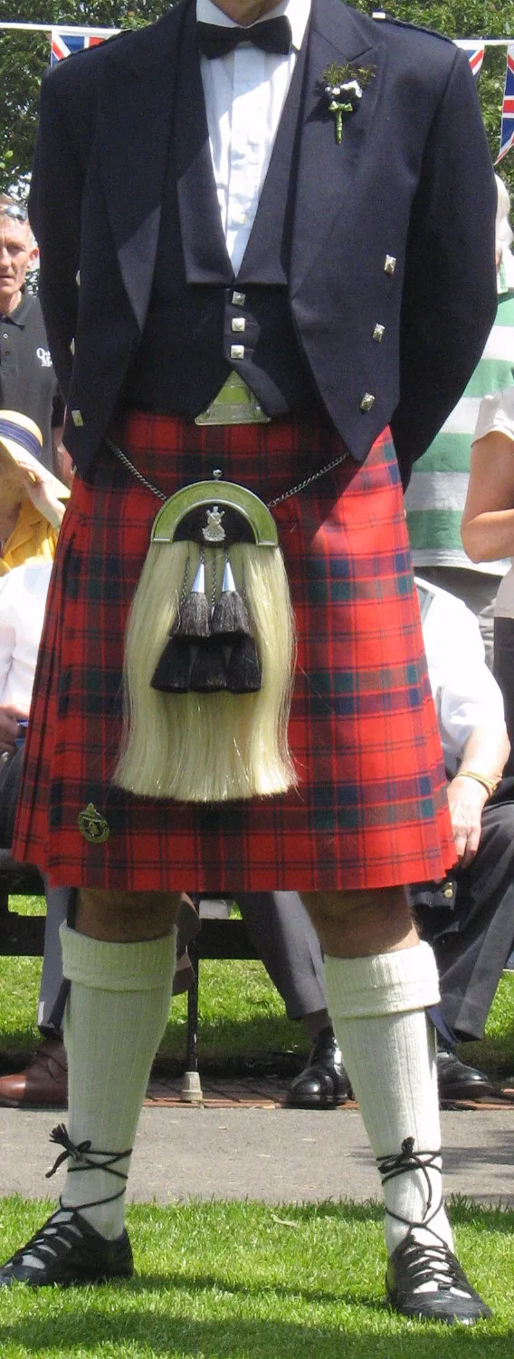 A man in a red tartan kilt with a horsehair sporran, white hose and a dark jacket, photographed from the chest down at an outdoor event