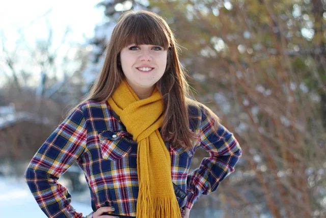 A smiling woman in a red, blue and yellow checked flannel shirt and a mustard scarf, standing outdoors in snow