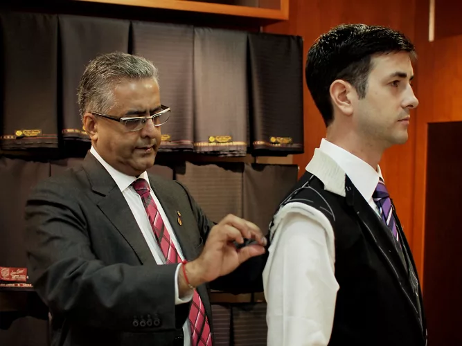 A tailor in a grey jacket takes measurements of a customer in a waistcoat in front of shelves of cloth